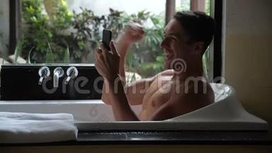 微笑的年轻人使用智能手机，而躺在浴缸在浴室。 在智能手机上得到好信息的人。 欢乐与欢乐